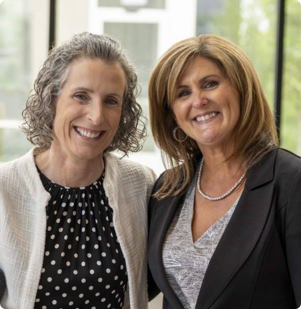 Two women smiling while standing side by side indoors. One has curly gray hair and wears a polka dot top with a light cardigan. The other has straight brown hair and wears a dark blazer over a patterned blouse. Theyre in front of large windows.