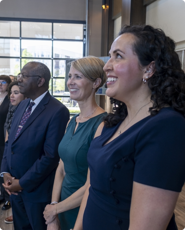 Three people, two women and one man, stand indoors in semi-formal attire, smiling and looking towards something off camera. Large windows and natural light are visible in the background.