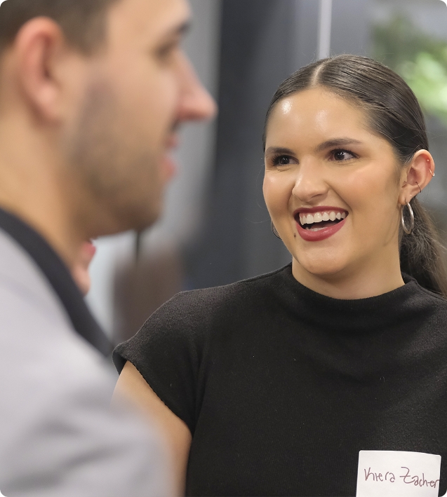 A person with dark hair and a black top is smiling and talking to another person in focus in the foreground. The setting appears to be an indoor, social or professional event.