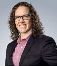 Person with curly brown hair and glasses, wearing a dark suit and a pink shirt, smiling against a gray background.