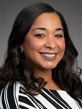A woman with long, wavy dark hair smiles at the camera. She is wearing a black and white striped top and has subtle makeup. The background is a plain gray.