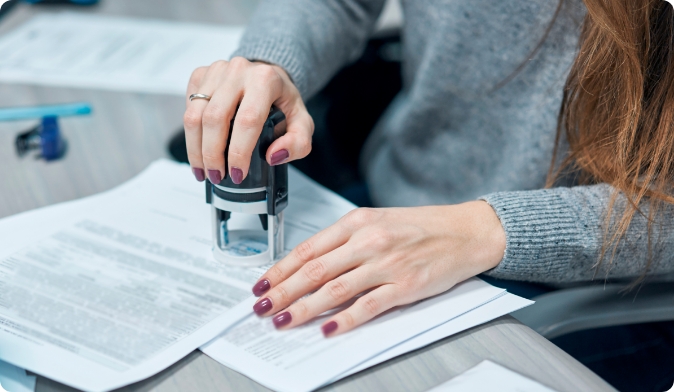 A person with long hair uses a stamp on a stack of documents placed on a desk. They are wearing a gray sweater, and their fingernails are painted dark purple. A pen lies nearby.