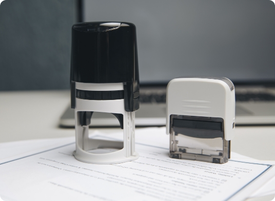 Two office stamps, one black and one white, are placed on a document with printed text. A laptop is in the background on a desk, slightly out of focus.