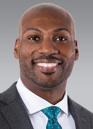 Smiling person wearing a gray suit, white shirt, and teal tie, with a plain background.