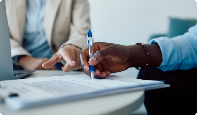 Two people are sitting at a table. One person is pointing at a document while the other holds a pen, poised to write. A laptop is visible on the table. The focus is on the hands and the document.