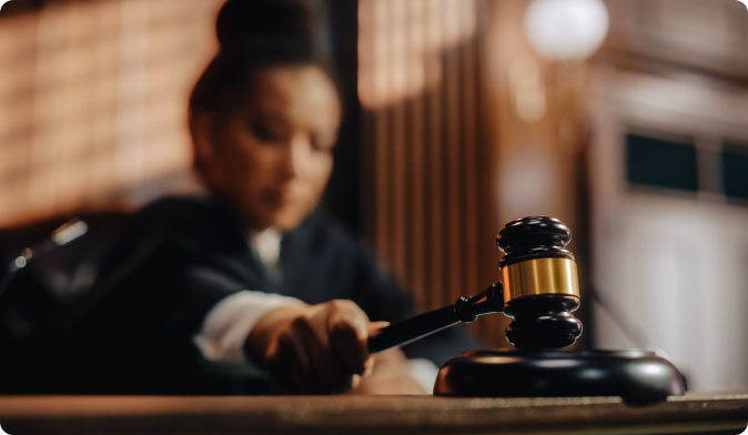 A judge in a courtroom is about to strike a gavel on a sound block. The scene is focused on the wooden gavel, while the background is softly blurred, highlighting the formal setting and the judges poised action.