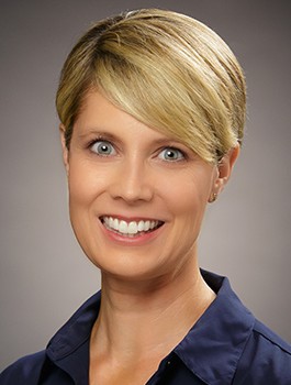 Smiling person with short blonde hair, wearing a dark blue collared shirt, against a neutral background.