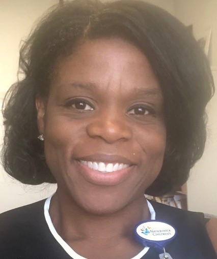 Smiling woman with short dark hair, wearing a black top with a white collar and a round name badge.