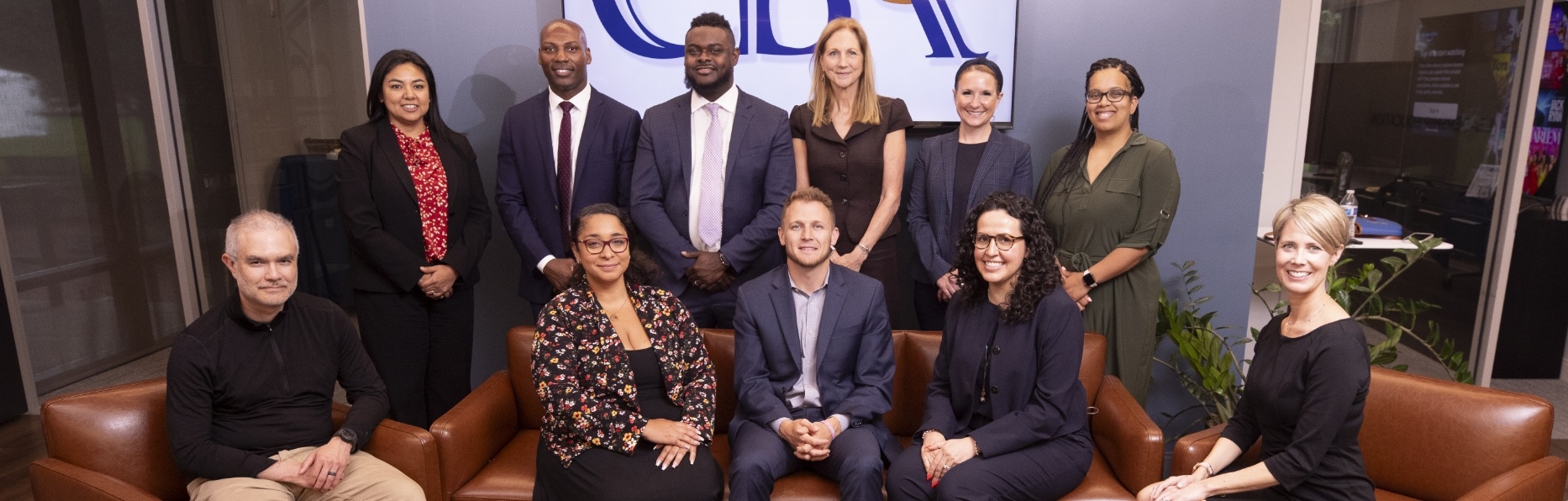 A group of eleven people, dressed in professional attire, pose for a photo in an office setting. Some are seated on a brown couch with others standing behind. A large sign with dark blue letters is visible in the background.