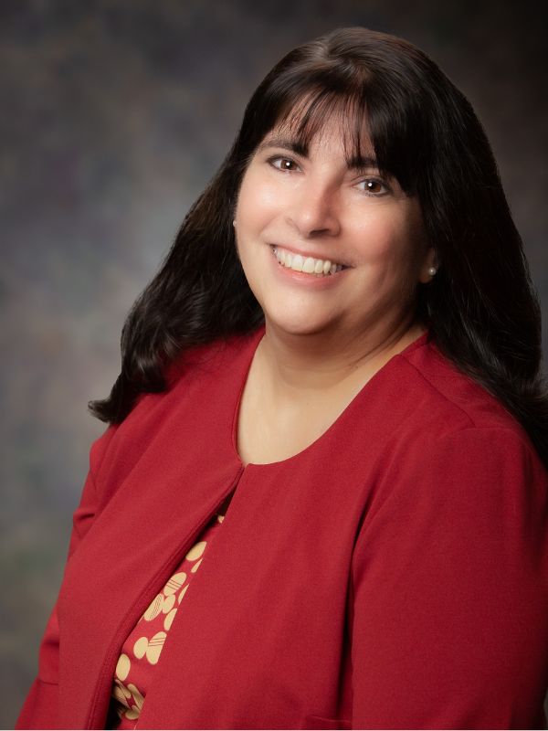 A woman with long dark hair, wearing a red blazer over a patterned top, smiles at the camera against a soft, blurred background.