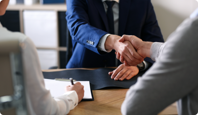 Two people shaking hands over a wooden table, one in a blue suit and the other in a gray sweater. A third person is taking notes on a clipboard. The setting suggests a professional meeting or interview in an office environment.
