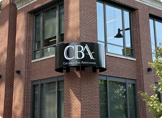 Sign on a brick building displaying CBA Columbus Bar Association in white letters with a gold scale of justice icon. Large windows are visible beneath the sign.
