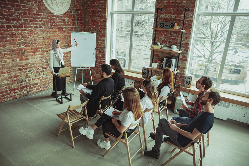A woman stands next to a whiteboard, addressing a group seated on folding chairs in a room with large windows and exposed brick walls. Some attendees take notes, and theres a laptop on a small table near the speaker.