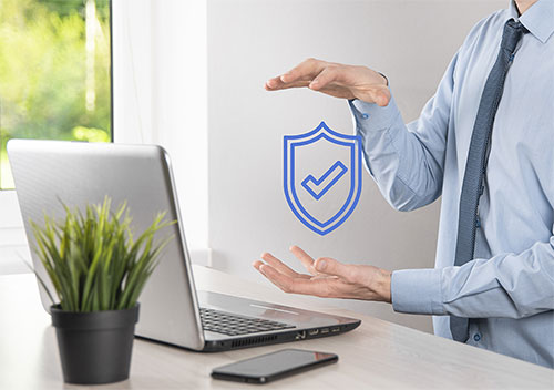 A person in a dress shirt gestures around a digital shield icon with a checkmark, symbolizing cybersecurity, near a laptop, smartphone, and potted plant on a desk.
