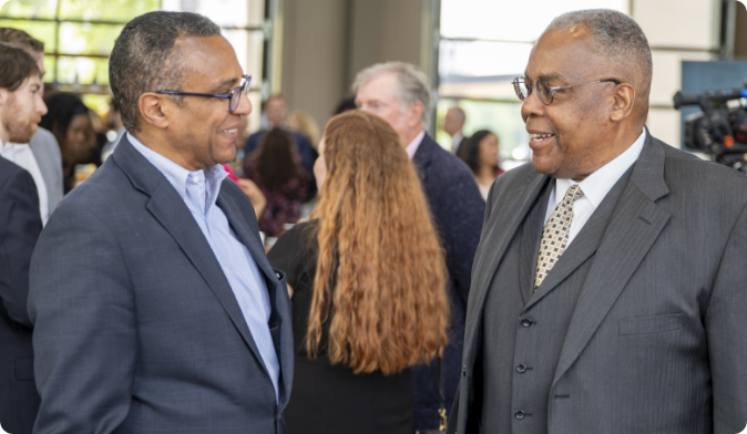 Two men in suits are smiling and conversing at an indoor event, with other people mingling in the background. The atmosphere appears professional and social.