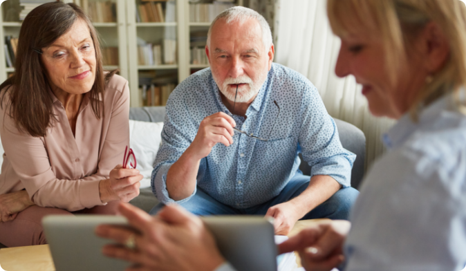Three people sit together in a cozy room, focused on a laptop screen. An older man with a beard and a woman with long hair appear attentive, while another woman gestures towards the screen, facilitating a discussion. Bookshelves are in the background.