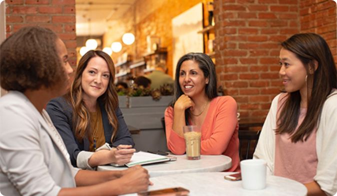 Four women sitting at a coffee shop table, engaged in conversation. One is holding a notepad. There are coffee cups on the table, and the setting has exposed brick walls and warm lighting in the background.