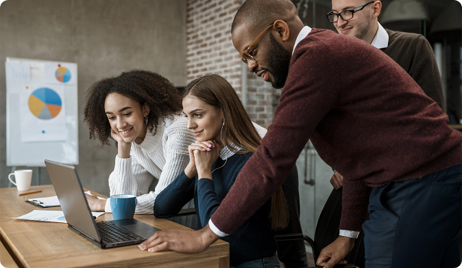 A diverse group of four people gathered around a laptop in an office, appearing engaged and smiling. A bar graph is visible on the wall in the background. A blue mug sits on the wooden table.