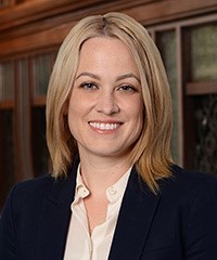 A woman with straight blonde hair, wearing a navy blazer over a white blouse, smiles at the camera in an indoor setting with wood paneling and glass behind her.