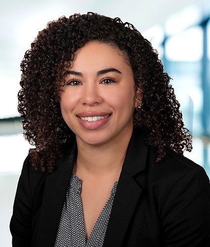 A woman with curly dark hair smiles at the camera. She is wearing a black blazer over a patterned blouse. The background is softly blurred, suggesting an indoor office setting.
