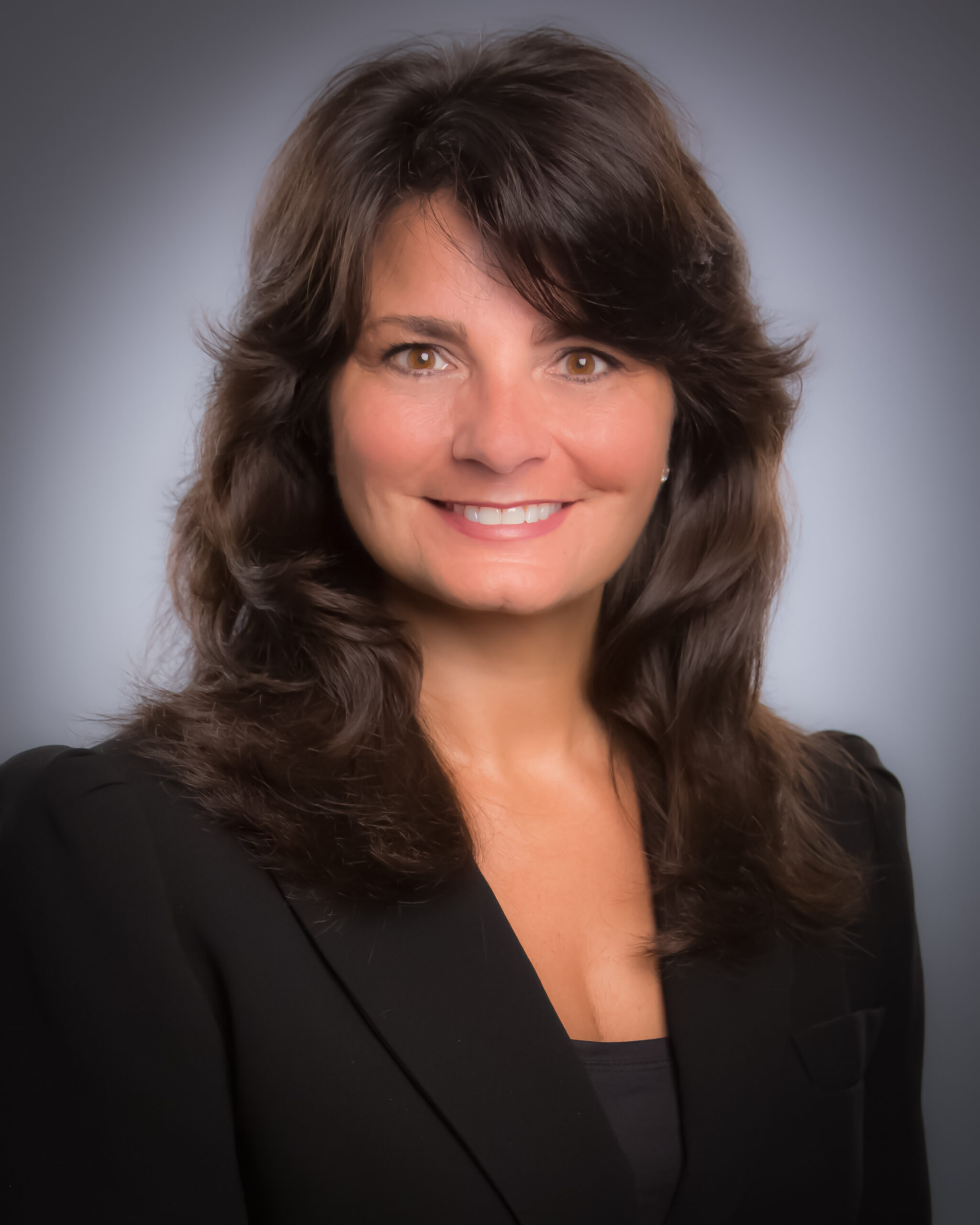 A woman with wavy dark brown hair wearing a black blazer and smiling, posing in front of a plain gray background.