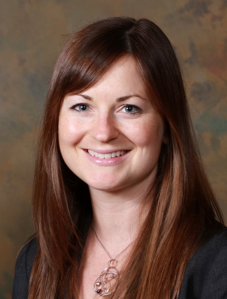 A woman with long, straight brown hair and blue eyes smiles at the camera. She is wearing a dark top and a silver necklace, with a brown and green mottled background behind her.