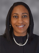A woman with straight dark hair wearing a black jacket, a pearl necklace, and hoop earrings, smiling in front of a gray background.
