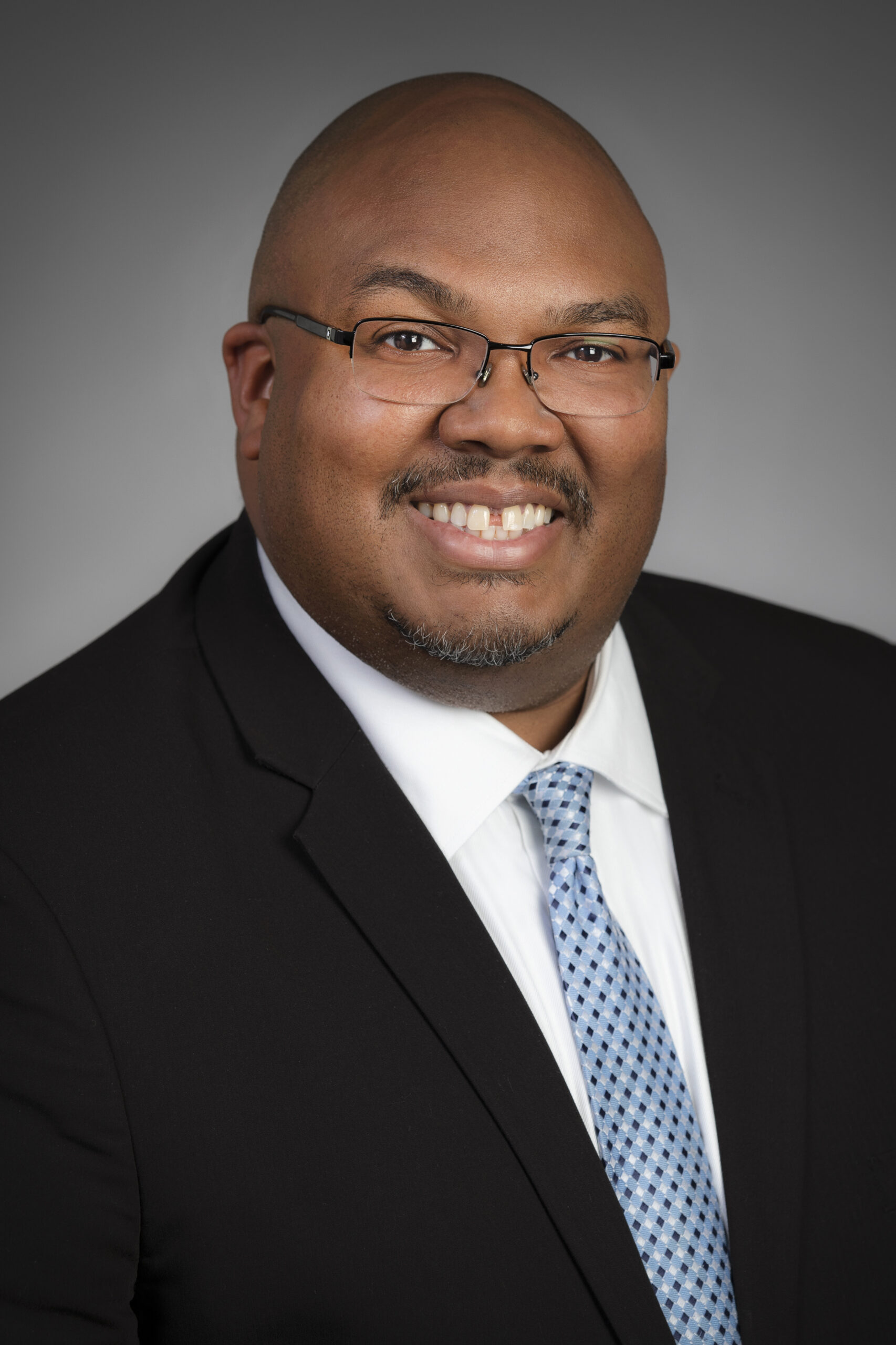A man wearing glasses, a black suit jacket, a white dress shirt, and a light blue patterned tie smiles at the camera against a plain gray background.