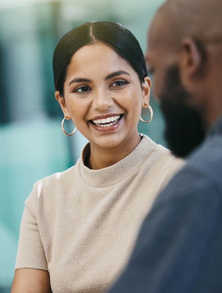 A woman with dark hair tied back, wearing hoop earrings and a beige top, smiles warmly while talking to a man whose face is only partially visible. The background is softly blurred.